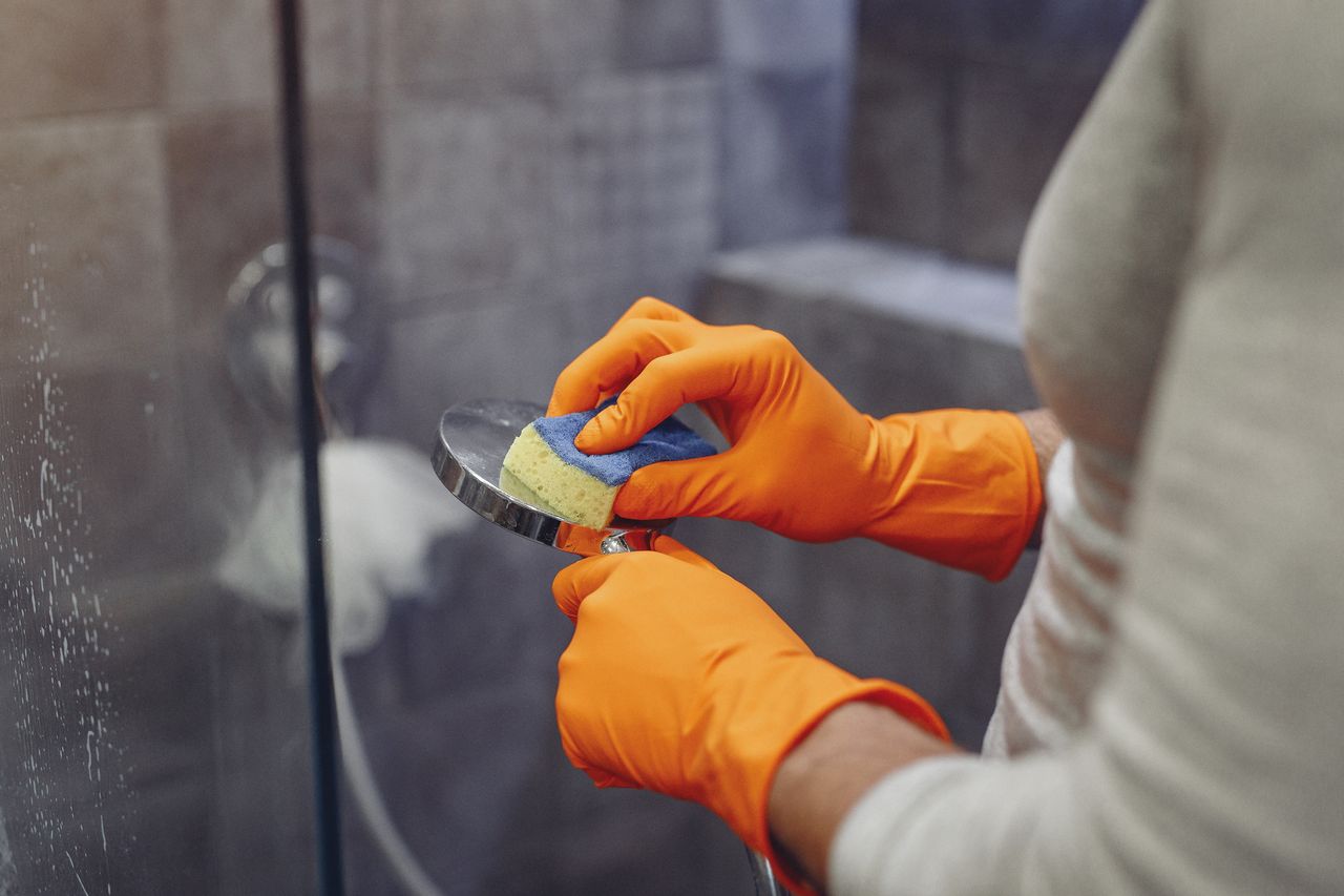 Close-up of a faceless man in orange rubber gloves wiping a shower nozzle with a sponge